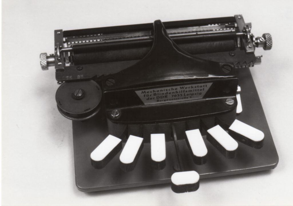 Vanha mekaaninen pistekirjoituskone. Siinä on kuusi valkoista näppäintä, jotka on asetettu puoliympyrän muotoon. Kirjoituskoneen yläosassa on metallinen sylinteri, joka toimii paperin syöttämiseen. Keskellä on metallinen laatta, jossa lukee saksaksi: "Mechanische Werkstatt für Blindenhilfsmittel der DDR 7035 Leipzig Burgauenstraße". Tämä viittaa siihen, että laite on valmistettu DDR:ssä, Leipzigissa.