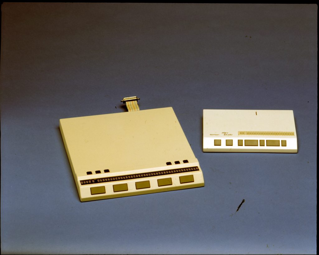 Kuvassa on kaksi vanhaa pistenäyttöä. Vasemmalla on suurempi laite, jossa on useita painikkeita ja pitkä rivi pistesuunnikkaita. Laitteen yläosasta lähtee litteä kaapeli. Oikealla on pienempi pistenäyttö, jossa on myös painikkeita ja braille-sunnkkaita. Pienemmän laitteen yläosassa lukee "Norman Micro Braille". Molemmat laitteet ovat vaalean värisiä ja ne ovat asetettu sinertävän harmaalle taustalle.