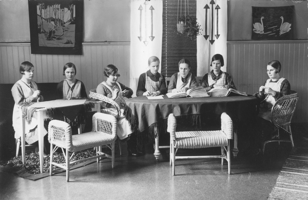 Mustavalkoinen valokuva, jossa seitsemän nuorta naista istuu huoneessa. He ovat pukeutuneet vanhanaikaisiin mekkoihin ja esiliinoihin. Kolme heistä istuu vasemmalla puolella olevan pöydän ääressä, ja neljä muuta istuu pyöreän pöydän ympärillä. Kaksi naisista neuloo, kun taas muut näyttävät lukevan tai kirjoittavan. Huoneen seinillä on kaksi taulua, joista toisessa on joutsenia. Huoneessa on myös rottinkituoleja ja -penkkejä sekä kasvi, joka roikkuu katosta. Taustalla on verhot, joissa on nuolikuvioita.