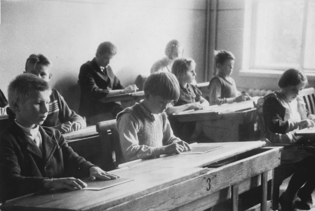 Mustavalkoinen valokuva esittää luokkahuonetta, jossa useat lapset istuvat vanhanaikaisissa puisissa pulpeteissa. Jokaisella lapsella on edessään pistekirjoitustaulu ja pistin, joita he käyttävät kirjoittaakseen pistekirjoitusta. Heidän sormensa liikkuvat keskittyneesti taulun päällä, kun he kirjoittavat tai lukevat. Luokkahuone on yksinkertainen ja valoisa, ja luonnonvalo tulvii sisään suuresta ikkunasta, joka sijaitsee huoneen oikealla puolella. Lasten vaatetus on yksinkertaista ja käytännöllistä, mikä viittaa siihen, että kuva on otettu menneiltä vuosikymmeniltä. Pulpettien etuosassa on numeroita, ja etualalla näkyy numero "3". Ympäristö on rauhallinen ja keskittynyt, mikä korostaa oppimisen ja keskittymisen ilmapiiriä.