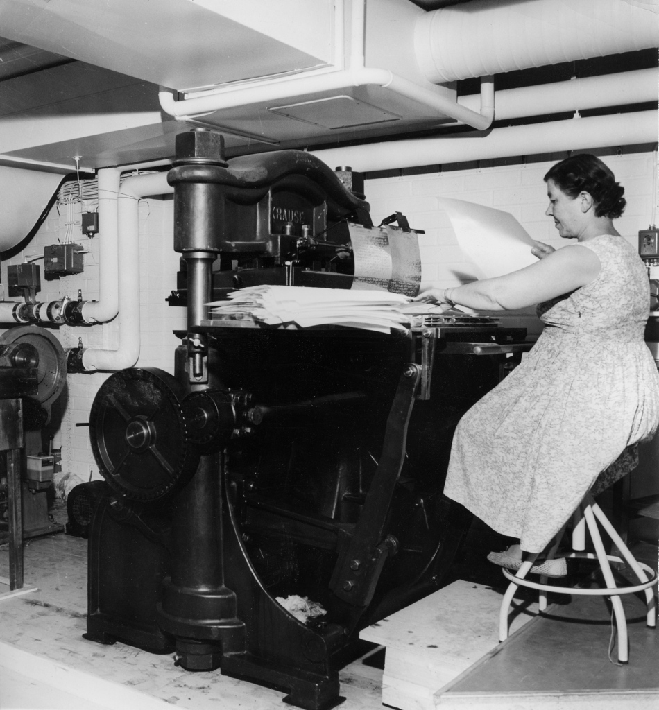 Nainen istuu jakkaralla suuren, vanhanaikaisen painokoneen ääressä. Hänellä on yllään kukkakuvioinen mekko ja hän käsittelee paperiarkkeja, joita on pinottu koneen päälle. Painokone on massiivinen, metallinen laite, jossa on suuria pyöriä ja vipuja. Taustalla näkyy valkoiseksi maalattu tiiliseinä ja putkistoa, joka kulkee katon rajassa. Ympäristö vaikuttaa olevan teollinen tai työpajamainen tila.