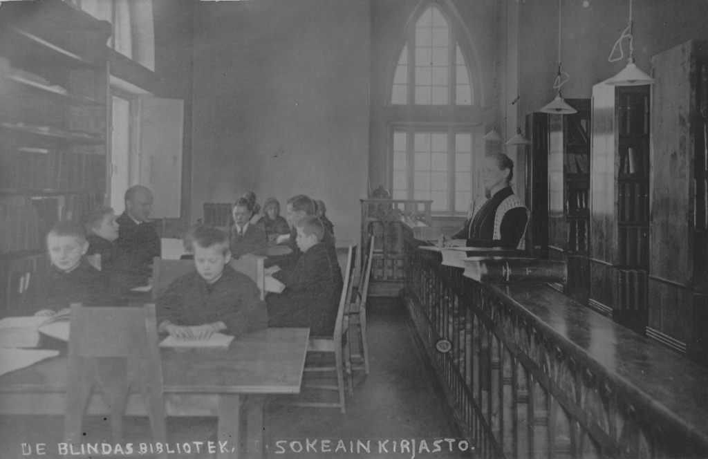 Vanha mustavalkoinen valokuva esittää kirjastoa, jossa on useita ihmisiä. Kuvan etualalla on kaksi poikaa, jotka istuvat pöydän ääressä ja lukevat kirjoja. Taustalla on useampia henkilöitä, jotka näyttävät myös olevan keskittyneitä lukemiseen tai keskusteluun. Oikealla puolella seisoo nainen, joka näyttää olevan kirjastonhoitaja tai opettaja. Huoneessa on korkeat ikkunat, jotka päästävät sisään luonnonvaloa. Seinillä on kirjahyllyjä, joissa on kirjoja. Kuvan alareunassa on teksti "DE BLINDAS BIBLIOTEK. SOKEAIN KIRJASTO."