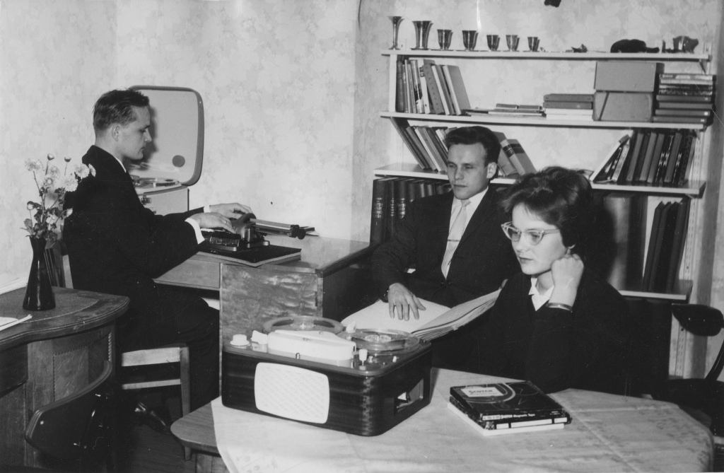 Mustavalkoinen valokuva, jossa on kolme henkilöä sisätiloissa. Vasemmalla puolella istuu mies, joka käyttää näkevien kirjoituskonetta. Hänellä on yllään tumma puku ja valkoinen paita. Keskellä istuu toinen mies, joka katsoo suoraan eteenpäin, ja hänellä on myös tumma puku ja valkoinen paita. Hänen toinen kätensä on pistekirjalla. Oikealla puolella on nainen, jolla on silmälasit ja hän katsoo alaspäin, ehkä lukien jotain. Hänen edessään pöydällä on nauhuri ja muutama kirja tai lehtiö. Taustalla on kirjahylly, jossa on kirjoja ja koriste-esineitä. Huoneen seinät ovat vaaleat ja niissä on kukkakuvioinen tapetti.