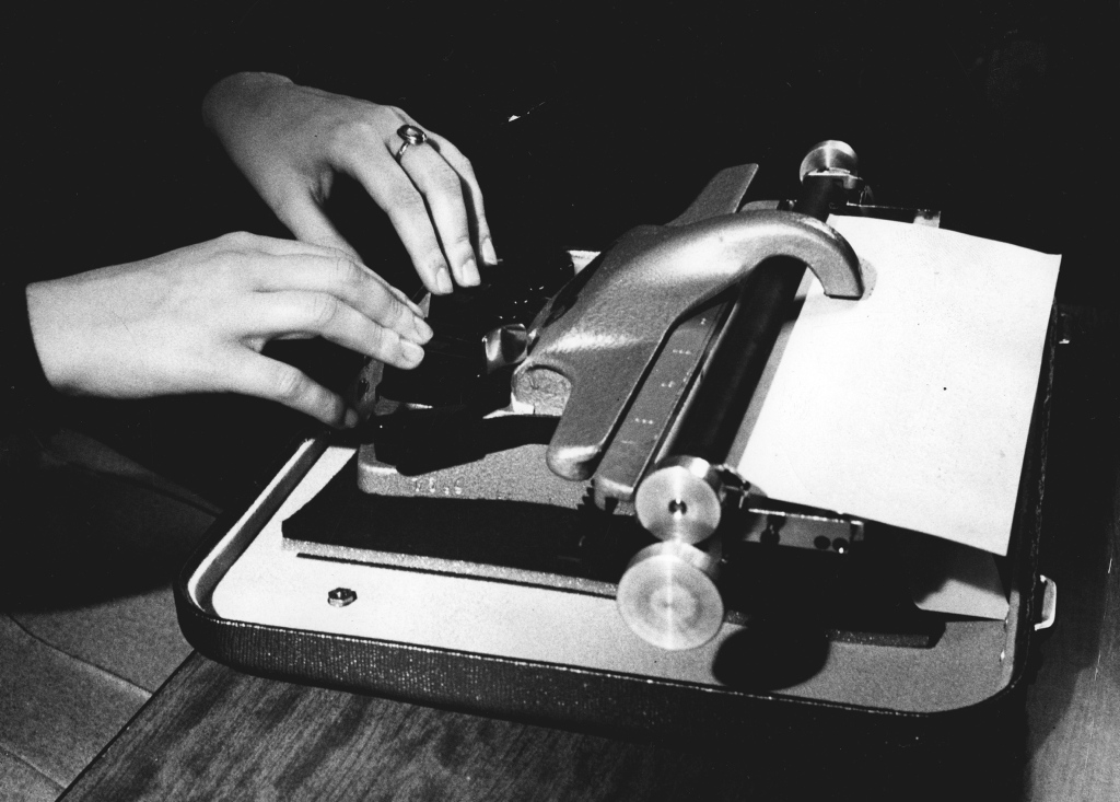 Kuvassa on mustavalkoinen otos, jossa näkyy Perkinsin pistekirjoituskone. Kone on asetettu pöydälle, ja siinä on metallinen runko. Koneessa on kuusi koholla olevaa näppäintä, jotka on suunniteltu pistekirjoituksen kirjoittamiseen, sekä välilyöntinäppäin. Koneen päällä on suuri kaareva vipu. Kaksi kättä on asetettu näppäimille, ja henkilön oikeassa kädessä on sormus. Koneessa on paperi valmiina kirjoittamista varten. Kuvassa ei näy koneen merkkiä, mutta se on tunnistettavissa Perkinsin pistekirjoituskoneeksi. Tausta on tumma, mikä korostaa koneen ja käsien yksityiskohtia.