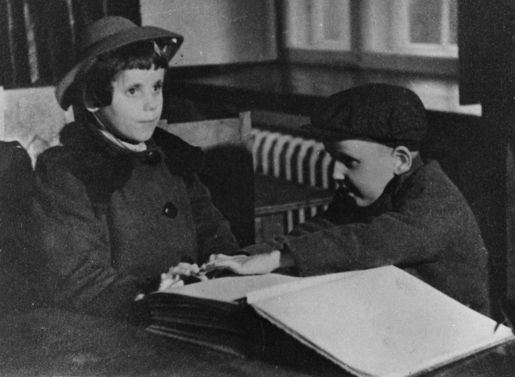 Vanha mustavalkoinen valokuva, jossa on kaksi lasta istumassa pöydän ääressä. Vasemmalla puolella on tyttö, jolla on lyhyet hiukset ja hattu päässään. Hänellä on yllään takki, jossa on suuret napit. Oikealla puolella on poika, joka on kumartunut eteenpäin ja koskettaa sormillaan kirjaa, joka on avoinna pöydällä. Kirja näyttää olevan pistekirjoituksella kirjoitettu. Poika käyttää lakkia ja hänellä on myös takki päällään. Taustalla näkyy patteri ja ikkunoita. Lapset näyttävät keskittyneiltä ja vakavilta, ja poika on syventynyt lukemiseen.