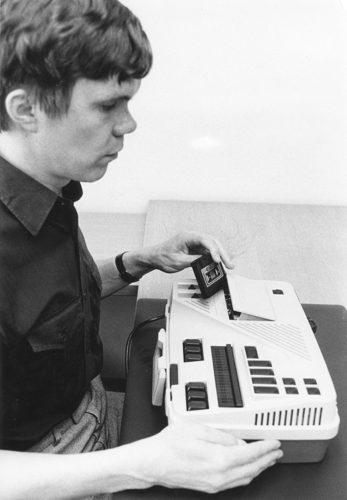 Mies istuu pöydän ääressä ja käyttää Versa Braille-laitetta. Hän on asettamassa kasettia laitteen sisään. Versa Braille on pöydällä hänen edessään, ja siinä on useita painikkeita ja vipuja. Miehellä on yllään tumma paita, ja hän näyttää keskittyneeltä tehtäväänsä. Kuva on mustavalkoinen, mikä antaa sille retrohenkisen ilmeen.