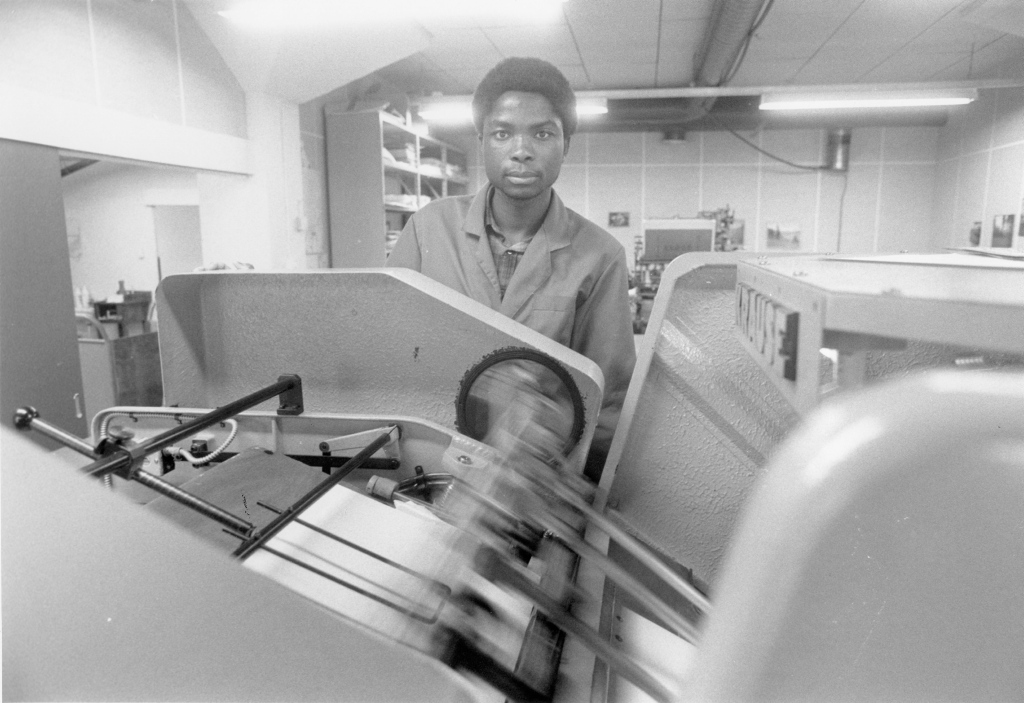 Mustavalkoinen valokuva teollisesta ympäristöstä. Kuvassa on nuori mies, jolla on lyhyt kihara tukka ja joka katsoo suoraan kameraan. Hänellä on yllään työtakki. Mies seisoo suuren koneen vieressä, joka näyttää olevan jonkinlainen painokone tai vastaava laite. Koneen osat liikkuvat nopeasti, mikä luo liike-epäterävyyttä kuvaan. Taustalla näkyy hyllyjä, joissa on erilaisia esineitä, sekä putkia ja johtoja katon rajassa. Ympäristö vaikuttaa olevan työpaja tai tehdas.