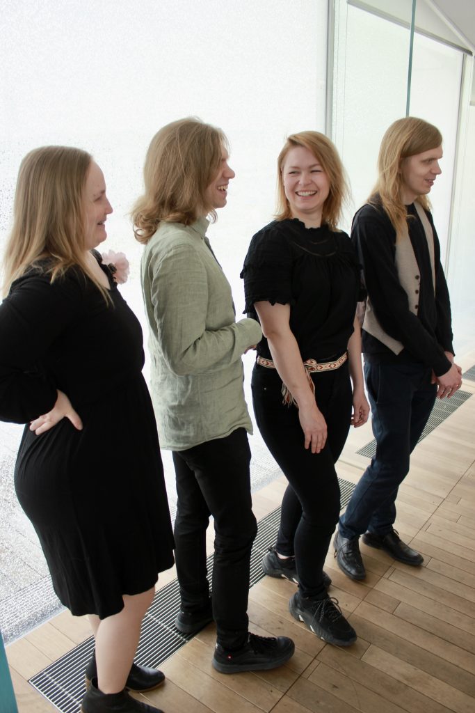 Neljä aikuista henkilöä seisomassa vierekkäin rivissä sisätiloissa, vaalean ikkunaseinän edessä. Kaikki katsovat ja hymyilevät tai nauravat oikealle päin, kohti jotakin tai jotakuta kuvan ulkopuolella. He näyttävät olevan iloisia ja hyväntuulisia.
• 
Vasemmalla on vaaleahiuksinen nainen, jolla on pitkät hiukset auki. Hänellä on päällään musta mekko tai tunika, jossa on koristeena vaaleanpunainen kukkaneula rinnassa.
• 
Hänen vieressään seisoo vaaleahiuksinen mies, jolla on hiukset sidottu niskaan pienelle poninhännälle. Hänellä on yllään vaaleanvihreä, pellavakankaalta näyttävä paita.
• 
Kolmantena on vaaleahiuksinen nainen, jolla on lyhyemmät hiukset. Hänellä on musta pusero, ja vyötäröllä värikäs, perinteisen näköinen kangasvyö solmittuna eteen.
• 
Oikeanpuolimmaisena seisoo vaaleahiuksinen mies, jolla on pitkät hiukset. Hänellä on musta paita ja vaalea, ruudullinen liivi. Kädet ovat rennosti housujen taskuissa.
Tausta on kirkas ja valoisa, suurista ikkunoista tulee luonnonvaloa sisään. Tunnelma on rento ja lämmin.