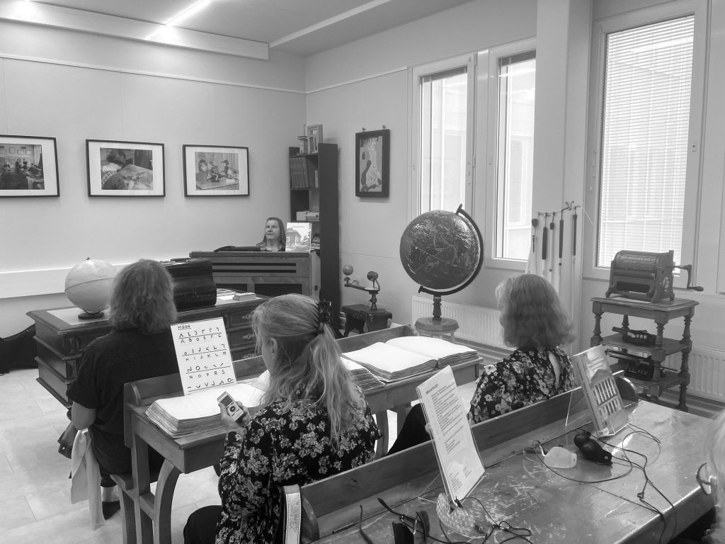 Kuva on mustavalkoinen ja esittää sisätilaa, joka näyttää vanhanaikaiselta luokkahuoneelta tai museohuoneelta.
Etualalla on kolme henkilöä istumassa puisissa pulpeteissa, selät kameraan päin. He lukevat suuria, kohokirjoituksella painettuja kirjoja, todennäköisesti pistekirjoja. Yhdellä pulpetilla on esillä myös pieni paperiarkki, jossa on kohokirjaimia. Keskellä kuvaa, hieman oikealla, näkyy pyöreä maapallo sekä suurempi taivaanpallo tukeilla.
Huoneen takaosassa, keskellä, istuu nainen vanhan urkuharmoonin ääressä. Hän katsoo eteenpäin, ehkäpä opettaen tai soittaen.
Seinillä on useita kehystettyjä valokuvia, jotka esittävät henkilöitä eri tilanteissa – ilmeisesti opetustilanteista. Yhdessä kuvassa näkyy esimerkiksi henkilö lukemassa pistekirjoitusta.
Oikealla, ikkunan alla, on vanhanaikainen kirjoituskone tai pistekirjoituslaite pienellä pöydällä.
Tunnelma on rauhallinen ja opettavainen, aivan kuin kyseessä olisi historiallinen opetushetki näkövammaisten koulussa tai sen museohuoneessa.