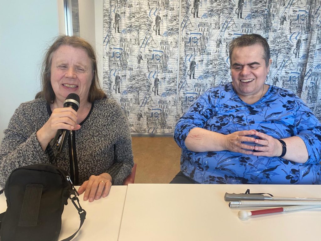 Kaksi aikuista naista istuu vierekkäin pöydän ääressä sisätilassa. Tunnelma vaikuttaa rennolta ja iloiselta.
Vasemmalla oleva nainen pitää kädessään mikrofonia ja näyttää puhuvan siihen. Hänen silmänsä ovat kiinni ja kasvoilla on keskittynyt mutta miellyttävä ilme, ikään kuin hän nauttisi hetkestä. Hänellä on vaaleanruskeat, hieman hartioille ulottuvat hiukset ja päällään harmaa, pehmeän näköinen neule. Pöydällä hänen edessään on musta laukku.
Oikealla oleva nainen istuu hieman taaksepäin nojaten ja hymyilee leveästi. Hän näyttää kuuntelevan vasemmanpuoleista naista ja nauttivan tilanteesta. Hänellä on lyhyet hiukset ja päällään sininen paita, jossa on tumma kukkakuvio. Hänen kädet ovat yhdessä edessä, ja pöydällä hänen puolellaan on valkoinen keppi sekä toinen samanlainen apuväline.
Taustalla on vaalea verho, jossa on kuvioita, kuten rakennuksia ja ihmishahmoja. Vasemmassa reunassa näkyy ikkuna, josta tulee luonnonvaloa. Alt-teksti on luotu tekoälyllä.