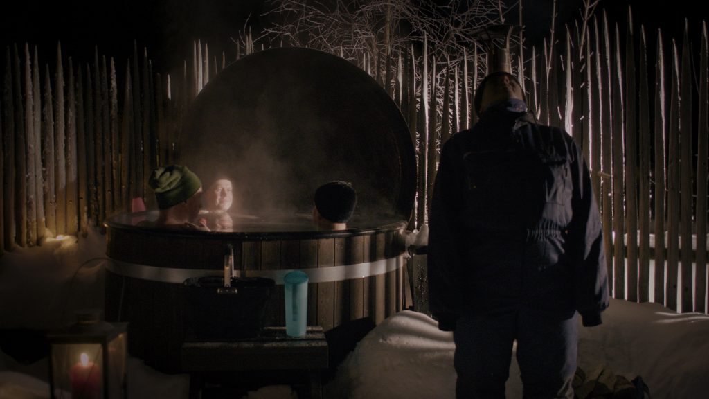 Valokuvan vasemalla puolella on palju, jossa on kolme alastonta hahmoa pipot päässä. Yhden hahmon kasvot ovat meihin päin. Kuvan oikealla puolella, etualalla on talvihaalariin ja pipoon pukeutunut Anu, joka on kääntänyt katseensa kohti taivasta. Öisessä kuvassa paljusta nousee höyryä ja etualalla, vasemmalla palaa lyhty.