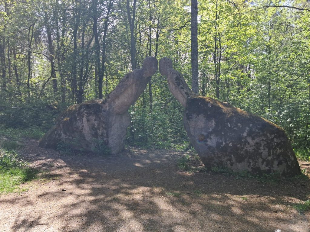 Metsäaukealla seisoo kookas kiviveistos, joka muodostuu kahdesta toisiaan vasten nojaavasta kivipaadesta. Kivet on muotoiltu muistuttamaan kahta ihmishahmoa, jotka kumartuvat hitaasti toisiaan kohti. Niiden yläosat kapenevat pyöreiksi "päiksi", jotka koskettavat kevyesti toisiaan otsasta tai kasvoista.
Hahmojen rungot ovat massiiviset ja kallistuvat etuviistoon. Ne näyttävät ikään kuin istuvan tai polvistuvan suurten kivijalkojen varassa. Kokonaisuus antaa vaikutelman hiljaisesta kohtaamisesta tai tervehdyksestä. Hahmot ovat lähes saman kokoiset, ja niiden asennot ovat peilikuvat toisistaan.
Kivien pinta on karkea ja epätasainen. Väri vaihtelee vaaleanharmaasta ruskehtavaan, ja pinnoilla kasvaa paikoin vihreää sammalta sekä vaaleita jäkälälaikkuja, jotka korostavat teoksen ikää ja luonnon vaikutusta.
Veistos sijaitsee metsän keskellä avoimella hiekkapohjaisella alueella. Taustalla kohoaa tiheä lehtimetsä, jonka joukossa kasvaa myös muutamia havupuita. Auringonvalo siivilöityy lehtien lomasta ja luo maahan pehmeän valon ja varjon verkoston. Tunnelma on rauhallinen ja lähes meditatiivinen.
Teoksen voimakkain vaikutelma syntyy siitä, että kaksi kivistä hahmoa näyttää olevan syvässä, sanattomassa yhteydessä toisiinsa – kuin ne vaihtaisivat hiljaisen tervehdyksen tai jakaisivat yhteisen hetken keskellä metsää. Alt-teksti on luotu tekoälyllä.