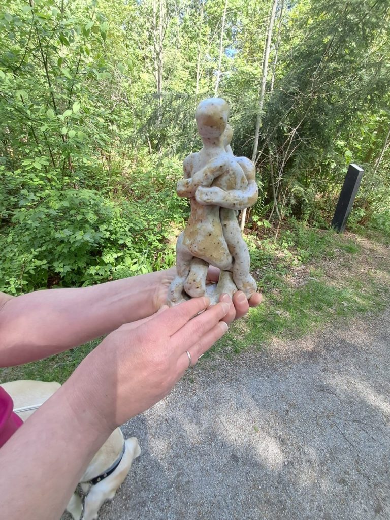 Henkilö pitelee molemmin käsin vaaleaa pienoismallia metsän reunassa kulkevan sorapolun varrella. Pienoismalli on noin kahden käden korkuinen, ja se esittää kahta toisiaan syleilevää tai tiiviisti toisiaan vasten seisovaa ihmishahmoa.
Hahmot ovat pelkistettyjä: päät ovat pyöreät ja kasvottomat, vartalot pehmeälinjaiset ja raajat paksut sekä hieman kömpelöt. Kasvonpiirteitä tai vaatteita ei ole kuvattu. Hahmot kietovat kätensä toistensa ympärille rinnan korkeudella. Ne seisovat jalat lomittain, mikä korostaa vaikutelmaa läheisyydestä ja yhteenkuuluvuudesta.
Veistos on valmistettu vaaleasta kivestä tai keraamisesta materiaalista. Sen pinnassa näkyy pieniä ruskeita pilkkuja ja epätasaisuuksia, jotka antavat sille luonnonkivimäisen ilmeen. Muotoilu on pehmeää ja pyöristettyä, eikä siinä ole teräviä kulmia.
Pienoismallia pitelevän henkilön sormet tukevat sitä alhaalta ja sivuilta. Vasemmassa nimettömässä näkyy sormus. Kuvan vasemmassa alakulmassa erottuu myös vaalean opaskoiran pää ja musta valjas, mutta koira näkyy vain osittain.
Taustalla levittäytyy vehreä metsä, jossa kasvaa lehti- ja havupuita. Oikealla näkyy tumma, pystysuora opastaulu tai infokyltti. Auringonvalo suodattuu puiden lomasta ja valaisee sekä pienoismallin että ympäröivän kasvillisuuden.
Kuva välittää vaikutelman tunnusteltavaksi tarkoitetusta pienoismallista, jonka avulla kävijä voi hahmottaa metsään sijoitetun suuremman veistoksen muodon käsin koskettelemalla ennen varsinaisen teoksen kohtaamista. Alt-teksti luotu tekoälyllä.