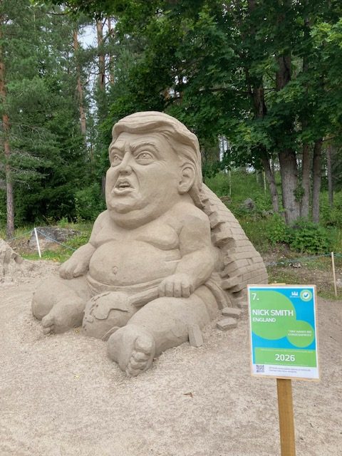 Suuri hiekkaveistos, joka esittää istuvaa, hyvin tunnistettavasti Donald Trumpia muistuttavaa mieshahmoa. Hahmo on veistetty tarkoituksellisen liioitelluksi ja karikatyyrimäiseksi.
Hahmo istuu maassa jalat levällään eteenpäin. Vartalo on paljas ja erittäin tanakka: suuri vatsa työntyy esiin ja rinnat, käsivarret sekä jalat on muotoiltu paksuiksi ja pyöreiksi. Kädet lepäävät reisien vierellä. Paljaat jalkaterät osoittavat eteenpäin, ja myös varpaat on veistetty näkyviin.
Kasvot ovat veistoksen hallitsevin osa. Pää on suuri suhteessa vartaloon. Suu on hieman auki ja huulet työntyvät eteenpäin. Katse suuntautuu ylöspäin ja sivulle. Posket ja leuan alue ovat voimakkaasti pyöristyneet. Hiukset on muotoiltu sivulle kammatuksi, Trumpille tyypillistä kampausta muistuttavaksi kokonaisuudeksi.
Hahmon selän takana kohoaa hiekkainen rakenne, joka muistuttaa osittain korkeaa tuolin selkänojaa tai seinämää.
Veistoksen oikealla puolella on vihreä-sininen esittelykyltti. Siinä lukee “7. NICK SMITH, ENGLAND” ja alhaalla 2026. Teoksen nimeksi näyttää olevan merkitty “THE EMPEROR'S NEW CLOTHES” eli Keisarin uudet vaatteet. Teoksen nimi selittää myös alastoman hahmon ja tekee veistoksesta poliittisen satiirin.
Veistos on ulkona hiekkaisella alueella. Taustalla kasvaa tiheää vihreää metsää, muun muassa mäntyjä ja lehtipuita. Veistosalue on rajattu ohuella köydellä. Kuvassa koko hiekkahahmo ja sen vieressä oleva esittelykyltti näkyvät lähes kokonaisuudessaan.