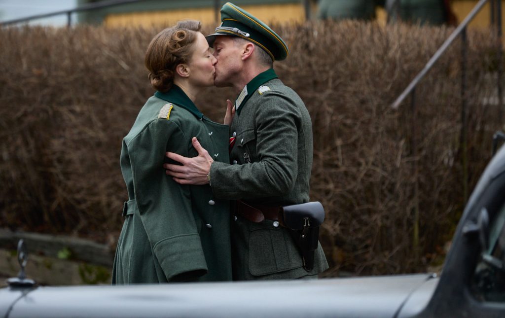 Saara (vas.) ja saksalaiseen upseerinunivormuun ja lippalakkiin pukeutunut Josef (oik.) suutelevat silmät kiinni. Saaran harteilla on vihreä sotilasmantteli, ja hänen vaaleanruskeat hiuksensa on kammattu kiharoille 1940-luvun tyyliin. Josefin vyöllä roikkuu pistoolikotelo. Taustalla on lehdetön pensasaita, ja etualalla näkyy vanhan harmaan auton konepelti.