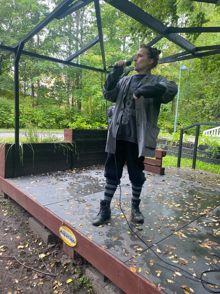 Ulkoilmalavalla esiintyy nuorehkolta vaikuttava, vaaleaihoinen henkilö, joka pitää oikeassa kädessään mikrofonia suunsa edessä. Hänen hiuksensa on nostettu ylös nutturalle, ja päälaella on aurinkolasit. Esiintyjällä on väljä harmaa takki, tumma painokuvioitu collegepaita, mustat housut, harmaa-mustaraidalliset polvisukat ja mustat maihinnousukengät. Vasen käsivarsi on koukistettuna rinnan korkeudelle voimakkaan eleen keskelle. Suu on avoinna, ja kasvoilla on keskittynyt, ilmeikäs olemus. Esiintyjä seisoo yksin matalalla, suorakulmaisella puulavalla, jonka tumma pinta on kostea ja syksyisten lehtien peitossa. Mikrofonin johto kiemurtelee lavan poikki. Lavan yläpuolella on mustista palkeista rakennettu avoin kehikko, ja ympärillä kasvaa tiheää, vielä pääosin vihreää lehtipuustoa. Kuva välittää vaikutelman intensiivisestä esityksestä rauhallisen vehreässä ulkoilmaympäristössä.