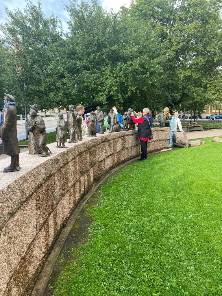Puistomaisessa ympäristössä kaartuu pitkä, matala, harmaista kiviharkoista rakennettu muuri, jonka päällä seisoo rivi erikokoisia pronssisia ihmishahmoja. Veistokset esittävät eri-ikäisiltä vaikuttavia, vanhanaikaisiin vaatteisiin pukeutuneita ihmisiä. Joukossa on muun muassa hattupäisiä aikuisia, pieniä lapsia sekä kumarassa asennossa oleva hahmo. Hahmot seisovat eri asennoissa ja katsovat eri suuntiin muodostaen ikään kuin menneiden aikojen ihmisjoukon. Osa veistoksista on lähes luonnollisen kokoisia, osa huomattavasti pienempiä. Muurin vierellä on useita ihmisiä tutustumassa teokseen. Yksi punahihaiseen asuun pukeutunut henkilö seisoo aivan muurin ääressä ja kohottaa kättään veistosten suuntaan. Toinen henkilö seisoo veistosten keskivaiheilla. Muurin sisäpuolella levittäytyy kirkkaanvihreä nurmikko, ja taustalla kohoaa suuria, tuuheita lehtipuita. Kivimuuri kaartuu kuvan vasemmasta etualasta kohti oikealla olevaa puistoaluetta. Pronssihahmojen tumma pinta erottuu vaaleasta kivestä ja vihreästä ympäristöstä. Kokonaisuus tuo mieleen eri sukupolvien ja aikakausien kohtaamisen.