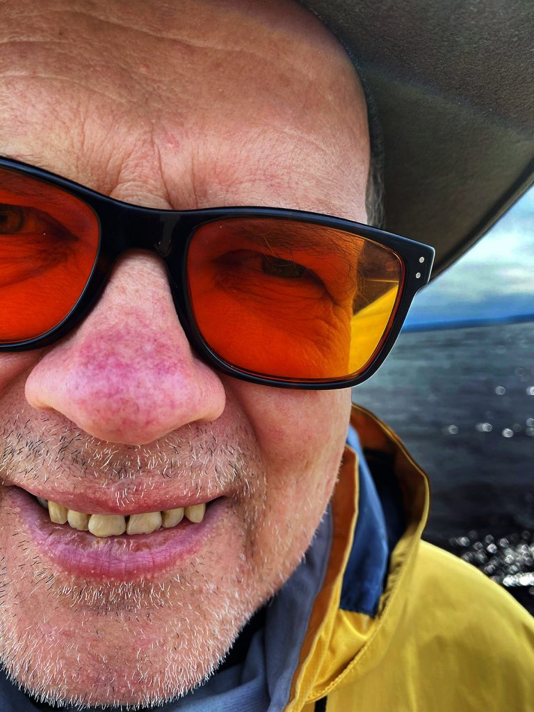 Pystysuuntainen lähikuva hymyilevästä miehestä ulkona veneessä. Kasvot täyttävät lähes koko kuvan, ja rajaus on hyvin läheltä otettu, hieman leikkisän selfiemäinen. Miehellä on mustakehyksiset aurinkolasit, joiden linssit ovat voimakkaan oranssit. Hän hymyilee leveästi niin, että hampaat näkyvät. Kasvoilla on vaaleaa sänkeä, ja nenä sekä posket ovat auringosta punoittavat.
Miehellä on kirkkaankeltainen takki, jonka alta näkyy tummansinistä vaatetta. Kuvan oikeassa reunassa hänen takanaan avautuu tumma, hieman kimalteleva vedenpinta ja kauempana matala horisontti sinisen taivaan alla. Yläreunassa näkyy veneen tummaa katosta tai kuomua. Kirkas auringonvalo korostaa kasvojen yksityiskohtia ja keltaista vaatetta. Kuvan tunnelma on aurinkoinen, välitön ja hyväntuulinen.