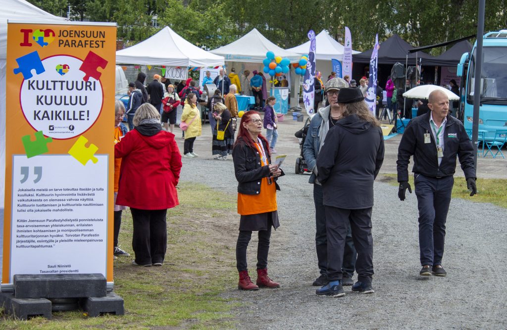 Vaakakuvassa Joensuun Parafestin ulkoilma-alueella liikkuu ja keskustelee runsaasti ihmisiä. Vasemmassa reunassa seisoo suuri oranssi tapahtumakyltti, jonka yläosassa lukee ”Joensuun Parafest” ja keskellä värikkäiden palapelinpalojen ympäröimänä ”Kulttuuri kuuluu kaikille!”. Kyltin alaosassa on tasavallan presidentti Sauli Niinistön tervehdys tapahtumalle.
Kyltin takana ja kuvan keskiosassa levittäytyy festivaalialue valkoisine tapahtumatelttoineen. Telttojen ympärillä on värikkäitä ilmapalloja, lippuja ja esittelypisteitä. Alueella seisoo ja kulkee eri-ikäisiä ihmisiä yksin ja pienissä ryhmissä. Osa keskustelee keskenään, osa tutustuu telttojen tarjontaan. Taustalla näkyy myös pyörätuolia käyttäviä osallistujia.
Kuvan etualalla punatukkainen nainen keskustelee kahden miehen kanssa. Oikealla tapahtuma-alueen laidassa kävelee mustiin pukeutunut mies vihreä henkilökorttinauha kaulassaan. Kuvan oikeasta reunasta näkyy osa suuresta turkoosista bussista.
Värikkäät tapahtumarakenteet ja ihmisten muodostamat pienet ryhmät tekevät näkymästä vilkkaan mutta rennon. Kuva välittää Parafestista avoimen, yhteisöllisen ja monenlaisia ihmisiä yhteen kokoavan kulttuuritapahtuman vaikutelman.